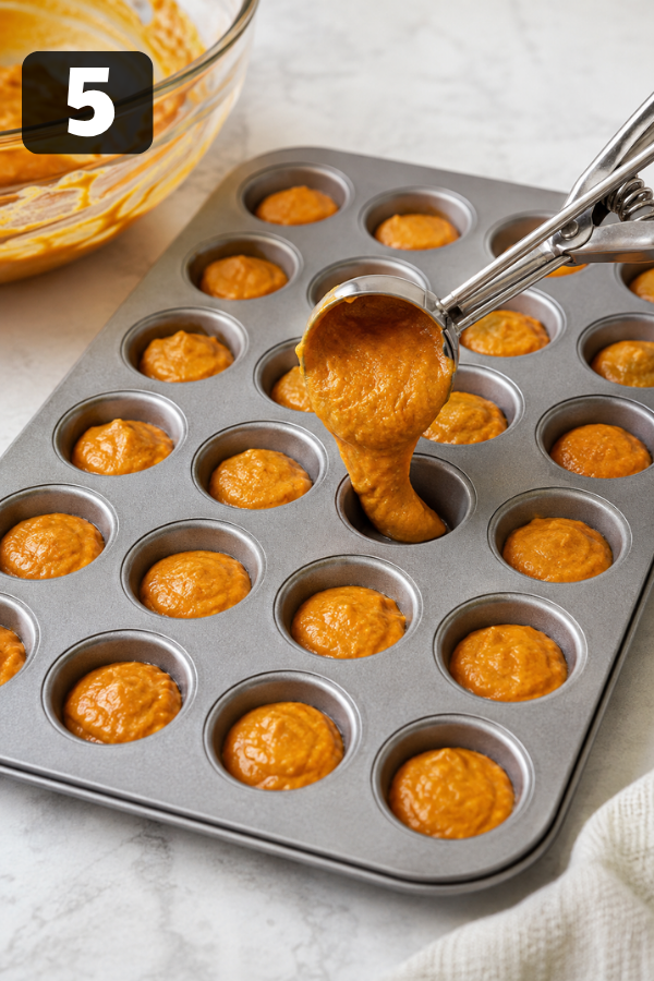 Pumpkin donut batter being scooped into a mini muffin pan before baking.
